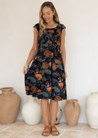 Model wearing a botanical print dress with scoop neck, cap sleeves and piped detailing at the waist and pockets.