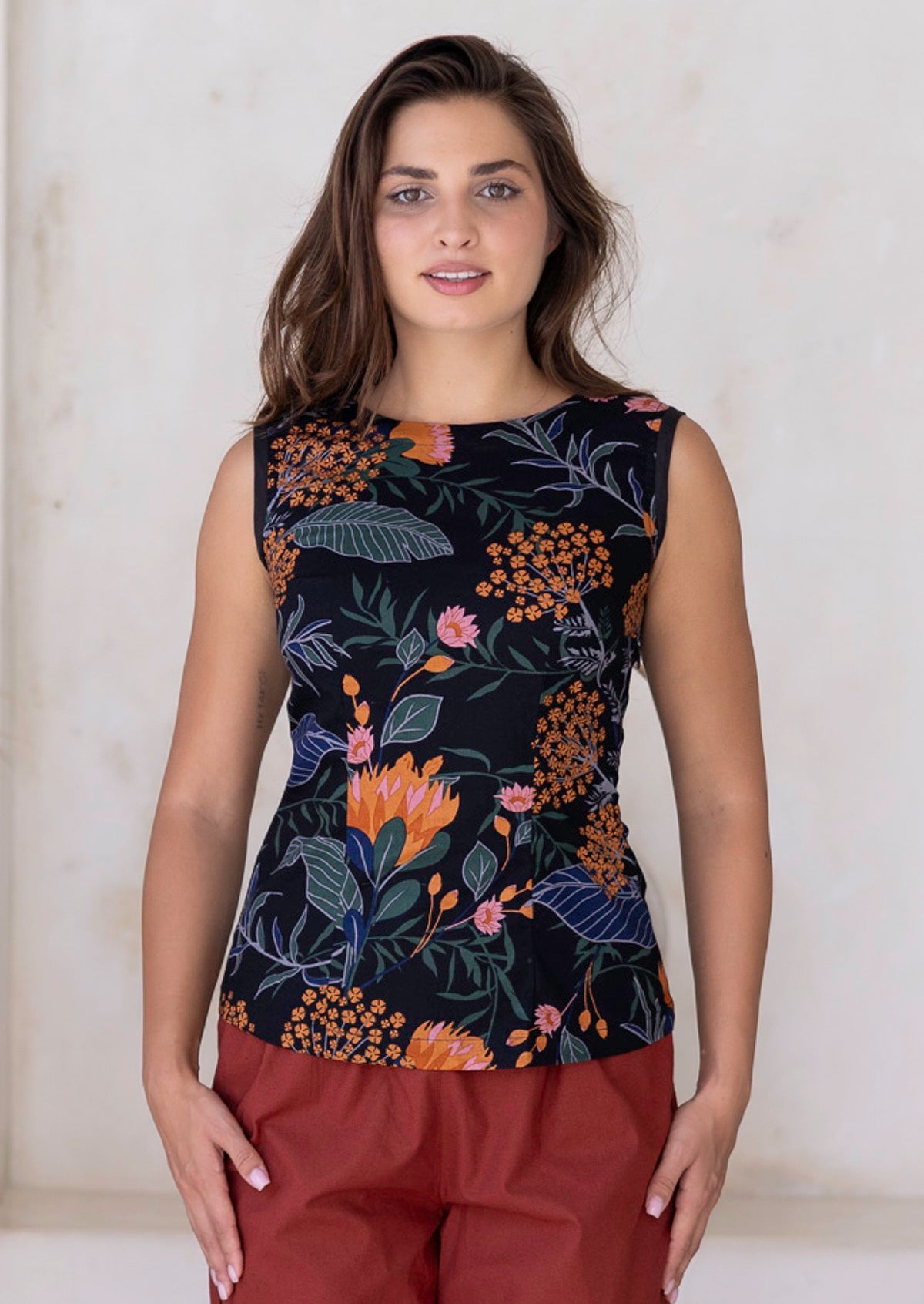 Woman wearing a sleeveless top with a black based floral print, paired with rust coloured pants 