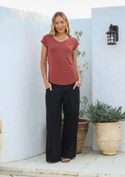 Woman wearing a 100% rayon terracotta V-neck top and black pants standing against a white wall with pot plants.