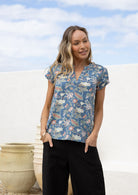Woman stands with hands in pocket wearing a sweet floral blouse featuring ruched shoulders and V neckline.