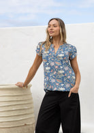 Woman posing in a dusty blue floral 100% cotton blouse with black cotton pants, standing against a white wall.
