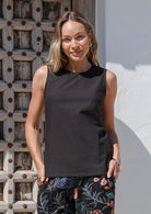 Woman wearing a lightweight 100% cotton black sleeveless top and floral pants standing against a wooden wall.