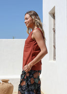Woman wearing a relaxed fit rust-colored sleeveless cotton top with side bust darts  and floral pants standing outdoors in front of white wall