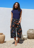 Woman stands with one hand in pocket wearing a black based botanical print wide leg, 3/4 length cotton pants with side zipper and pockets
