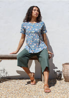 Woman sitting on a wooden bench wearing a floral print relaxed fit top and green pants against a white wall.
