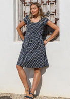 Woman wearing a 100% cotton knee length cap sleeved summer dress with round neckline and tiny decorative pleats from center of the neckline