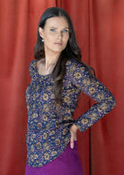 Woman standing sideways with hand in back pocket of skirt, wearing 100% cotton long sleeve top with tiny pleated details and round neck.