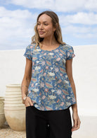 Woman wears dusty blue based floral cotton cap sleeve top with pin tuck detail across the bust and a scoop neckline