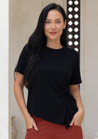 Woman wearing a black top with a knot detail with hands in pockets of rust pants