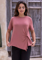 Woman wearing a pink t-shirt with a knot detail over black cotton pants in front of a rustic wooden fence.