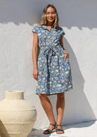Woman in a blue floral cotton dress with cap sleeves, buttoned bodice, pockets, box pleats at the waist and a fabric waist tie.