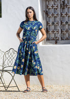 Woman wearing a blue floral cotton dress standing in front of a white wall with hands in her pockets