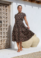 Model standing in skirt flare pose wearing a 100% cotton button through dress with maroon, cream florals on black base.