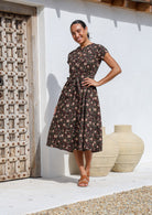Woman stands in cotton 50's style midi length dress with short sleeves, pockets and a detachable waist tie.