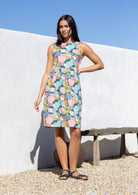 Woman wearing a pastel coloured print sleeveless dress standing against a white wall with blue sky.