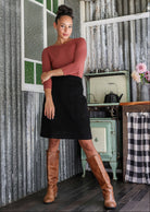 Woman in a cotton corduroy black A-line skirt paired with a dusty pink top and tan boots in a rustic kitchen