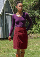 Woman wearing a corduroy skirt standing outdoors with a rustic building and greenery in the background.