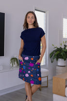 A woman standing in a modern room, wearing a knee-length skirt with a multicoloured spot print, with her hands in pocket