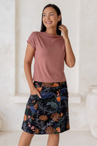 Woman stands in 100% cotton above knee length A-line skirt with piped detailing, pockets and a side zipper.