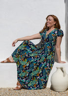 Woman wearing a cap-sleeve maxi dress with a colourful peacock print in teal, blue, yellow, and orange, sitting on a white stone ledge next to a white ceramic jug.
