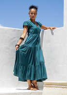 Woman in a teal maxi dress standing against a white wall with blue sky.