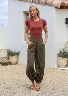Woman wearing a basic rayon top and olive green genie style cotton pants with cuffed ankles, standing outdoors with potted plants in the background.