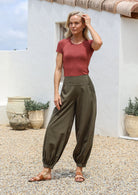 Woman standing outdoors wears 100% cotton relaxed fit pants in olive green with pockets, elastic at back of waist and cuffed ankles.