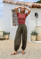 Woman posing outdoors in Aladdin style cotton pants in olive green with wide flat front waistband, pockets and buttoned ankles