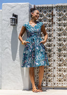 Model with both the hands in pocket wears 100% cotton dress with crossover bodice, cap sleeves and fuller skirt in teal based floral print.