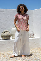 Woman wearing a rose pink based floral 100% cotton top and white pants standing in front of a decorative vases.