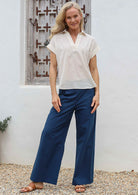 Woman standing in front of wooden window wears cotton shirt style collared top in natural white with v-neckline and folded short sleeves