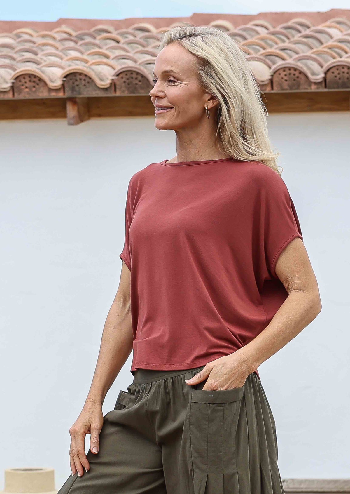 Woman smiling standing sideways with hand in pocket wearing 100% rayon batwing top in terracotta standing in front of a white house