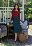 Woman in a red long sleeve top and green cotton corduroy A-line midi length skirt next to chairs and a table