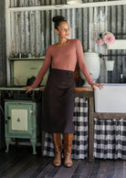 Woman standing in a rustic kitchen wearing a chocolate brown cotton corduroy A-line midi length skirt paired with a rose pink top