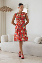 Woman standing in a living room wears knee length 100% cotton dress in vibrant red backdrop accented by elegant multicoloured floral vines.