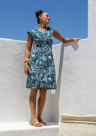 Woman standing in a living room wears knee length 100% cotton dress with delicate multicoloured wildflowers on a teal base.