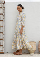 Woman stands side ways wearing a 100% cotton tiered maxi dress in neutral based floral print with 3/4 sleeves, pockets and mandarin collar