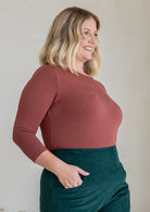 Woman standing sideways wearing a rayon boat neck terracotta pink 3/4 sleeve top.