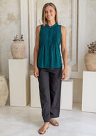 Woman wearing a teal sleeveless top with pleats across the bodice and dark pants standing in an indoor setting with decorative vases.