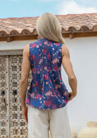 Woman wearing a blue floral sleeveless top with decorative pleats at the front and back, paired with white cotton pants, standing against a rustic door.