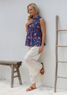 Woman wearing a floral sleeveless top with decorative pleats, high round neckline and white pants leaning against a white wall.