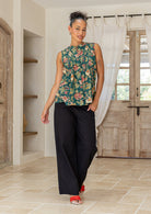 Woman wearing a floral sleeveless top with decorative pleats, high round neckline and black pants in a room setting.