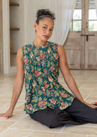 Woman sitting on a tiled floor wearing a green based floral sleeveless 100% cotton top and black pants.