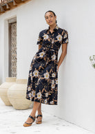 Woman wearing 100 percent cotton button through shirt dress with collar and short sleeves, in a black based floral print. 