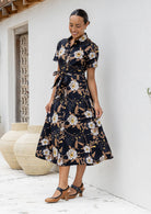 Model standing in front of white wall in floral printed shirt dress with detachable fabric belt, short sleeves and pockets.