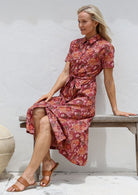 Woman sitting on a wooden bench wears 100% cotton shirt style dress with collar, pockets and button through front in warm toned floral print.