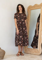 Woman standing next to a large mirror wears a floral black cotton dress with three tiered design, short sleeves and pockets