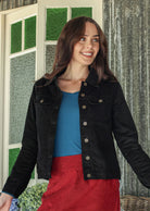 Woman wearing a black cotton corduroy jacket over a blue top and red skirt, standing in front of a window with green panes.