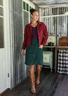 Woman wearing a corduroy jacket, green skirt, and brown boots standing in a rustic setting.