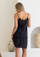 Black cotton cami slip dress on model, back view, in sunlit room with woven chair and pottery.